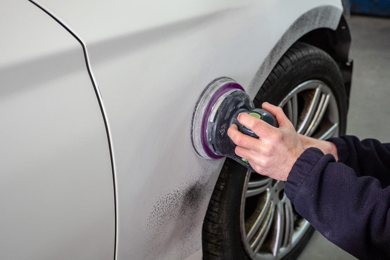 修理工場のスタッフが、電動サンダーを使用して白い車のボディ表面を研磨している様子。