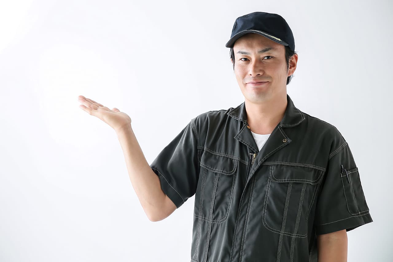 黒いつなぎと作業帽を着用し、笑顔で案内ポーズをとる男性整備士。