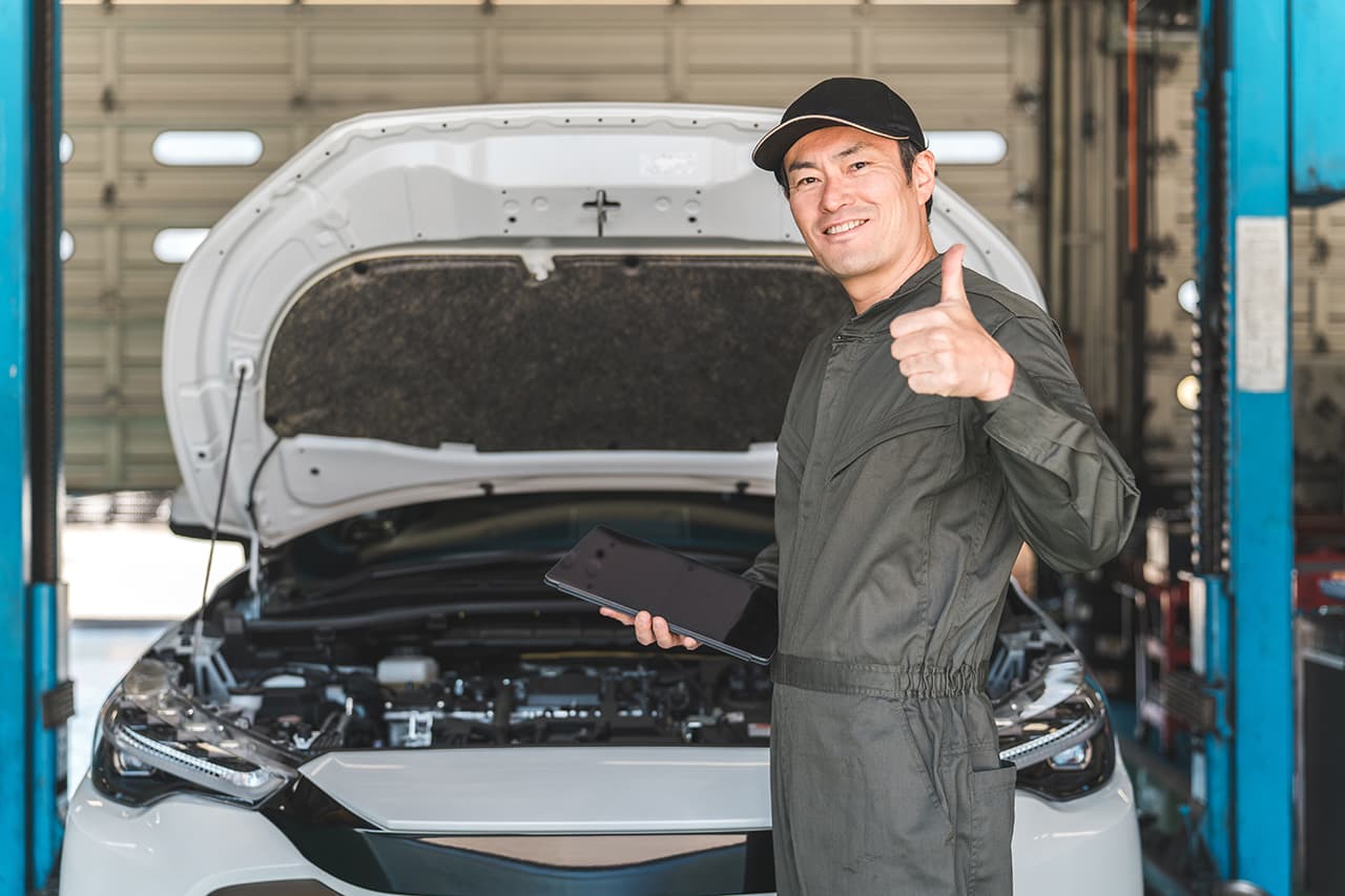 自動車整備工場で、ボンネットを開けた白い車の前に立ち、タブレットを手に笑顔の男性整備士