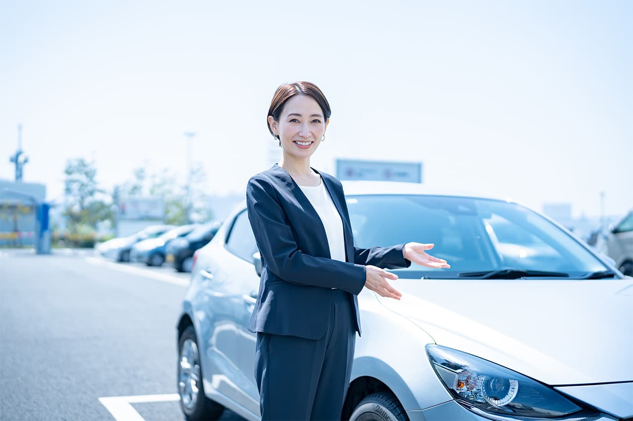 屋外駐車場でシルバーの乗用車の横に立ち、笑顔で案内ポーズをとるスーツ姿の女性スタッフ。