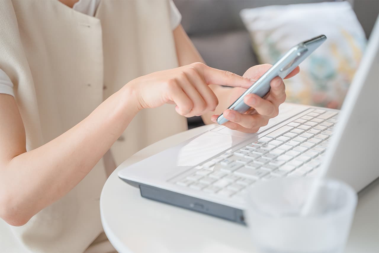 ノートパソコンの前でスマートフォンを操作する女性の手元